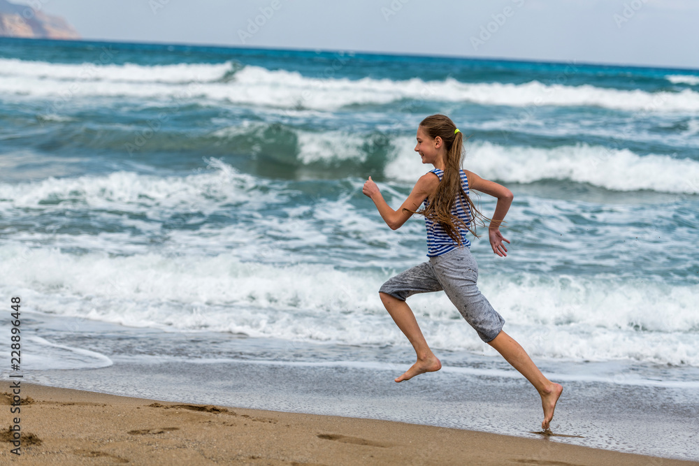 Cute happy Girl in striped t-shirt running along the beach in jumping over waves. Beautiful summer sunny day, blue sea, picturesque landscape. Cute happy Girl in striped t-shirt running along the beach in jumping over waves. Beautiful summer sunny day, blue sea, picturesque landscape.