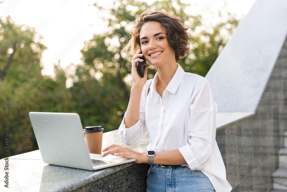 Cute beautiful woman standing outdoors using laptop computer talking by phone. Cute beautiful woman standing outdoors using laptop computer talking by phone.
