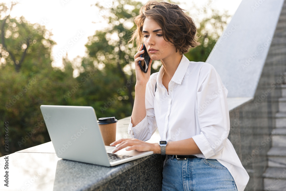 Obraz premium Displeased beautiful woman standing outdoors using laptop computer talking by phone.