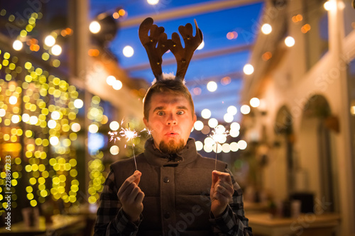 Christmas, people and winter holidays concept - surprised man in horns of deer standing at night street with bengal lights in his hands