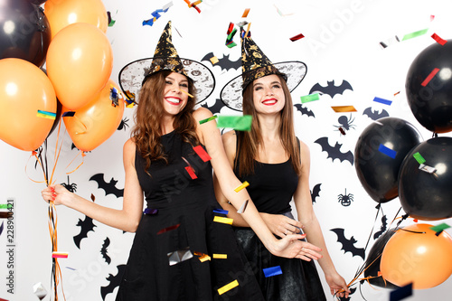 Two beautiful girls in black dresses and witch`s hats have fun with black and orange balloons and confetti.Halloween party