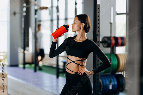Slim dark-haired girl dressed in black sportswear drinks water in the gym near the sport equipment