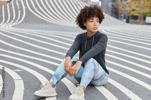 Outdoor shot of pensive dark skinned teenager dressed in casual sportsclothes, sneakers, sits on asphalt, recreats after having sport competitions, has fit body. Teenagers and active lifestyle concept photo