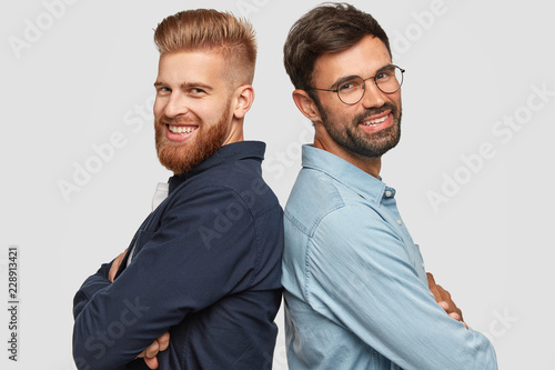 Best friend stand back to back, have friendly relationships, keep hands folded, smile gladfully, work as team, isolated over white background. Happy foxy bearded man and his partner pose indoor photo