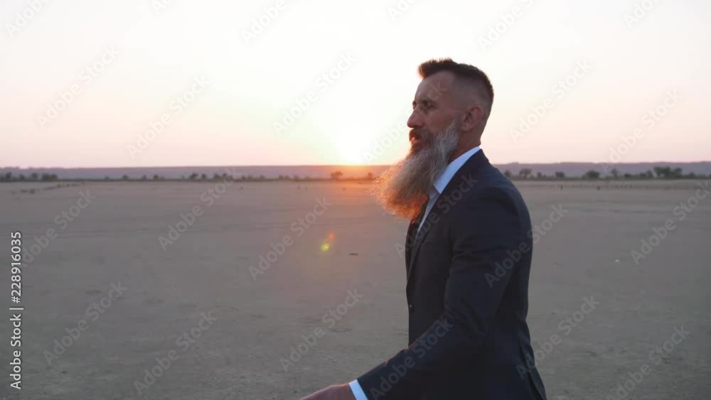 Handsome bearded mature man in suit walking outdoors on beach during sunset, slow motion Handsome bearded mature man in suit walking outdoors on beach during sunset, slow motion