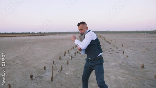 Mature handwome man in suit with long gray beard boxing with shadow outdoors on beach, slow motion photo