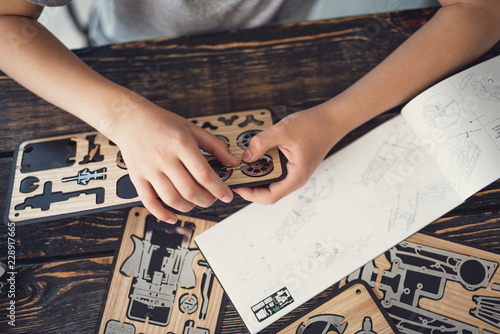 Curious kid sitting at the table and using the instructions while working with the wooden constructor