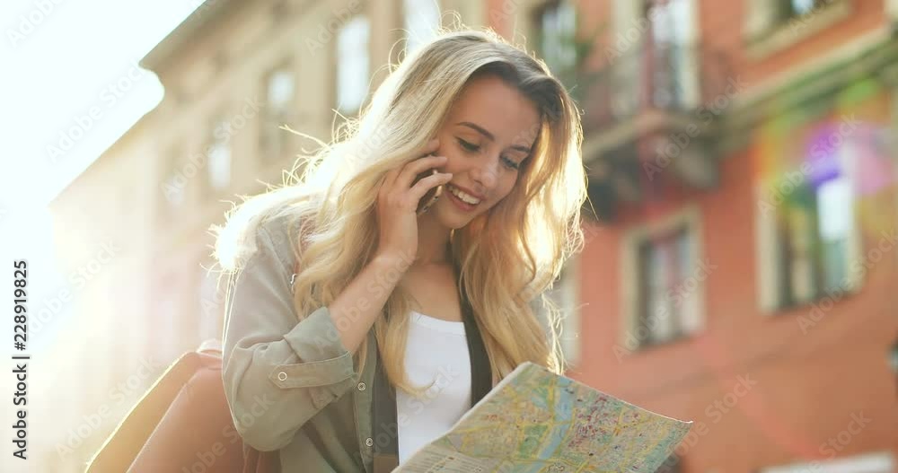 Portrait shot of the happy blonde young woman chatting on her phone while walking the city with a map in hands. Close up. Outdoors. Portrait shot of the happy blonde young woman chatting on her phone while walking the city with a map in hands. Close up. Outdoors.