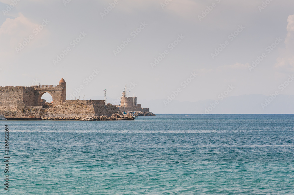 View at old fortification in old port from Akti Sachtouri sea promenade with Delphinia, sculpture of Dolphins.  Rhodes, Old Town, Island of Rhodes, Greece, Europe.