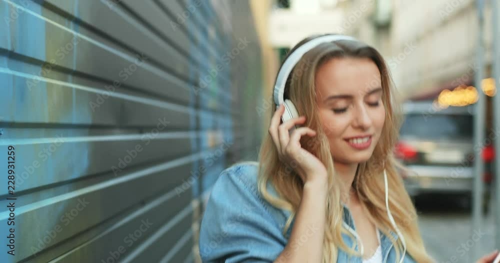 Beautiful blonde young woman in the headphones stepping the street and listening to the nice music on the smartphone. Close up. Beautiful blonde young woman in the headphones stepping the street and listening to the nice music on the smartphone. Close up.