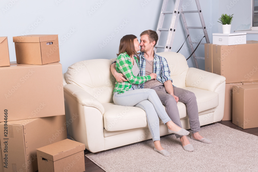 Moving, new apartment and relocation concept - young couple sitting and hugging on the couch surrounded with boxes Moving, new apartment and relocation concept - young couple sitting and hugging on the couch surrounded with boxes