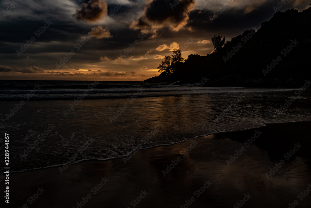 An amazing golden sunset behind a mountain silhouette edge on the end of Karon Beach, Phuket Thailand. A dramatic sky with vibrant light and reflections in the ocean