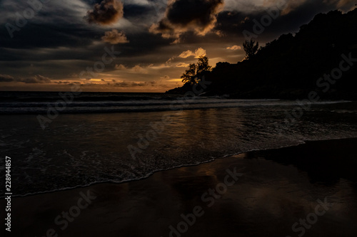 Obraz na płótnie An amazing golden sunset behind a mountain silhouette edge on the end of Karon Beach, Phuket Thailand. A dramatic sky with vibrant light and reflections in the ocean w salonie