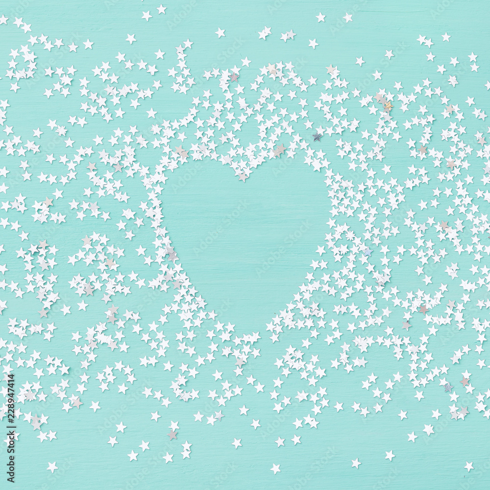 Closeup of small silver confetti stars in the shape of a heart on a turquoise background. The concept of the season of holidays