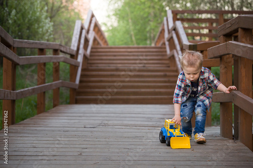 Cute boy is playing with a blue-and-yellow tractor in the park against the wooden stairs background.