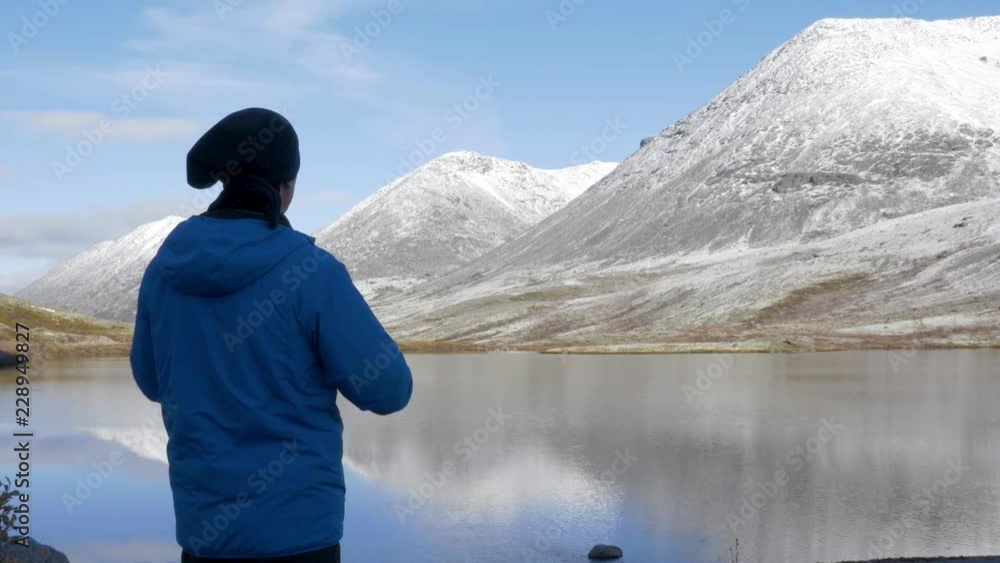 A man stands on a high point in the mountains and admires a beautiful view. There are peaks around it and a beautiful lake below. He photographs landscapes on a smartphone A man stands on a high point in the mountains and admires a beautiful view. There are peaks around it and a beautiful lake below. He photographs landscapes on a smartphone