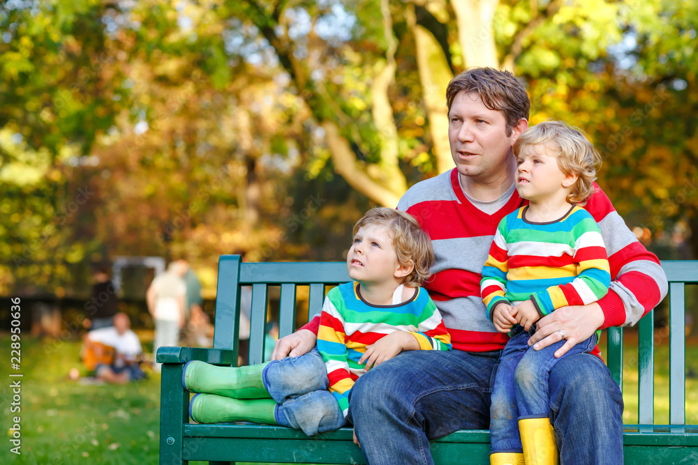 Fototapeta premium Two little kid boys and young father sitting together in colorful clothing on bench. Cute healthy children, siblings and their dad having fun in autumn park on warm sunny day. Happy family of three.
