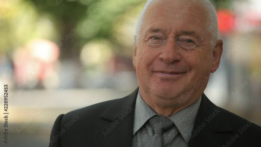 Cheery bokeh portrat of a smiling white-haired man in a neat business suit with a grey tie standing and smiling in a green park in autumn Cheery bokeh portrat of a smiling white-haired man in a neat business suit with a grey tie standing and smiling in a green park in autumn