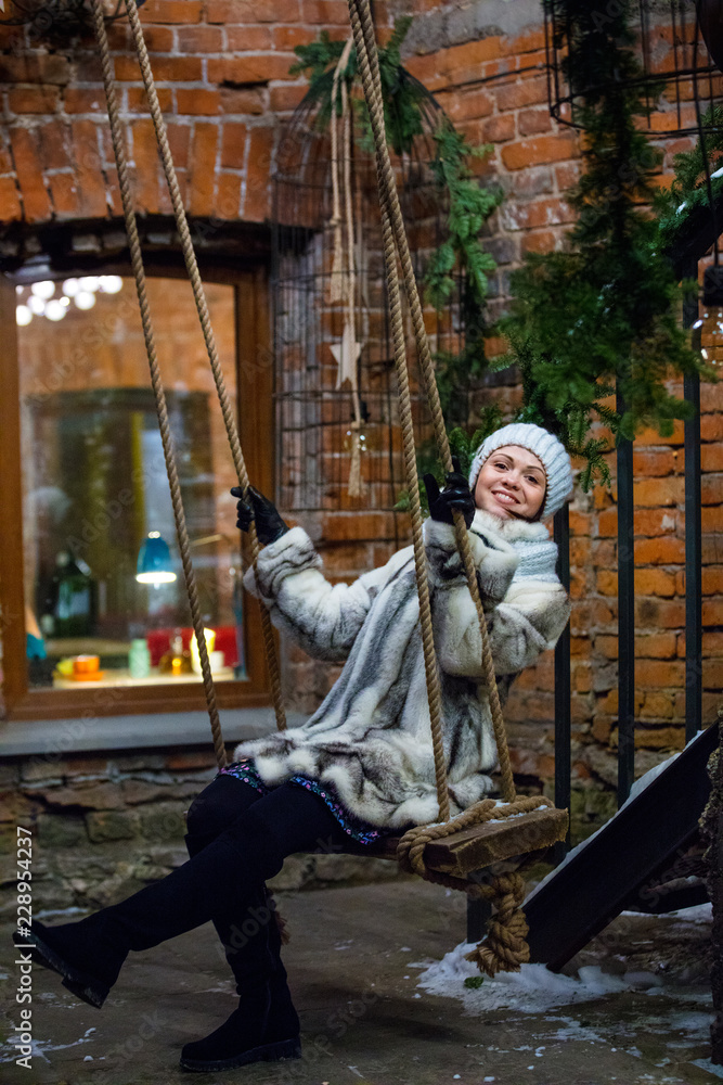 young woman in  fur coat, hat and snood on swing against window of cafe