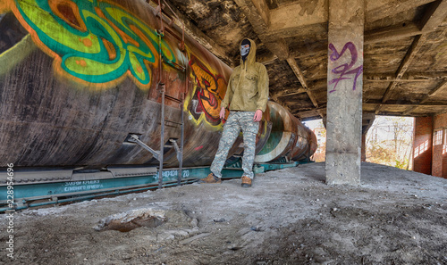 Bandit or terrorist in hood and balaclava mask staing ear the old rusty fuel tank train in abandoned ruined building photo