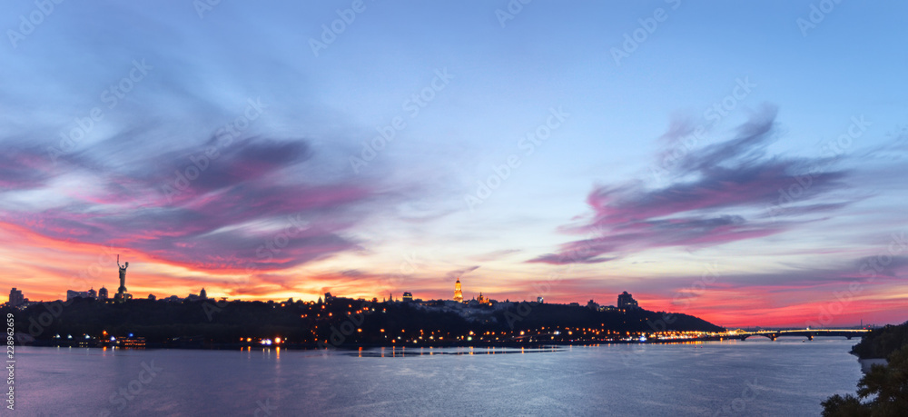 Sunset lit by colors, evening cityscape on the skyline of the monastery Lavra and Monument to Motherland. Kiev panoramic view with colorful clouds in the sky and reflections in the Dnieper river