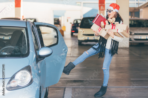 Young woman opening door of her car with leg (foot) while her hands are full of christmas gifts and presents boxes, after xmas shopping in mall photo