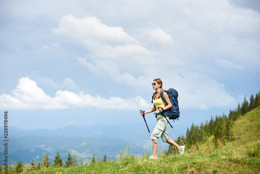 Attractive woman tourist hiking mountain trail, walking on grassy hill, wearing backpack and sunglasses, using trekking sticks, enjoying summer cloudy day. Outdoor activity, tourism concept Attractive woman tourist hiking mountain trail, walking on grassy hill, wearing backpack and sunglasses, using trekking sticks, enjoying summer cloudy day. Outdoor activity, tourism concept