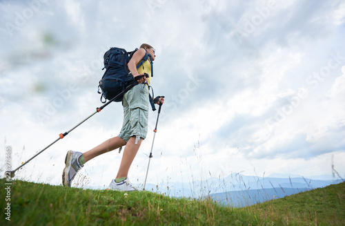 Low angle view of attractive woman hiker hiking in Carpathian mountain trail, walking on grassy hill, wearing backpack, using trekking sticks, enjoying summer day. Outdoor activity, lifestyle concept