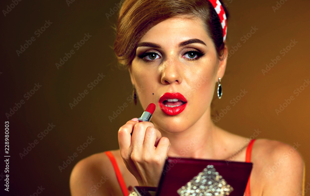 Girl in pin up retro style do makeup. Makeup with red lipstick. Sexy girl  look down and paints lips and looks in small mirror on gradation brown  background. Stock Photo | Adobe