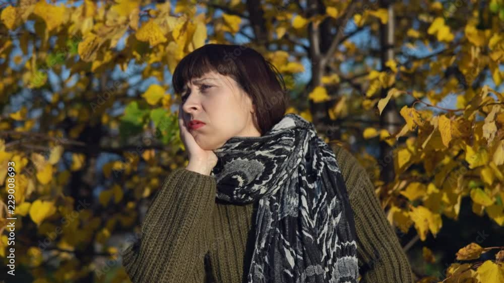 A young sick woman in a green knitted sweater strokes her head against the background of yellow foliage in the city park in Indian summer. A young sick woman in a green knitted sweater strokes her head against the background of yellow foliage in the city park in Indian summer.