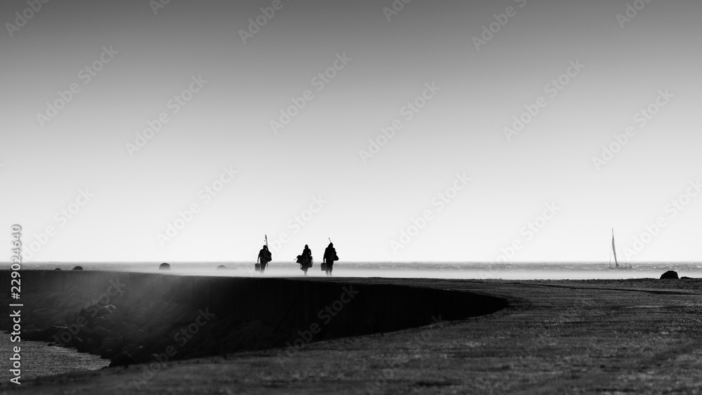 Silhouette of fishermen returning from their fishing spot in a concrete sea pier on a windy late afternoon in a sand cloud, Black and White Photo, Figueira da Foz, Portugal Silhouette of fishermen returning from their fishing spot in a concrete sea pier on a windy late afternoon in a sand cloud, Black and White Photo, Figueira da Foz, Portugal