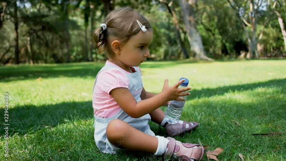 Female toddler opening bottle of water and sitting on green grass in park. Cute little child in sunny park in slow motion. Childhood concept Female toddler opening bottle of water and sitting on green grass in park. Cute little child in sunny park in slow motion. Childhood concept