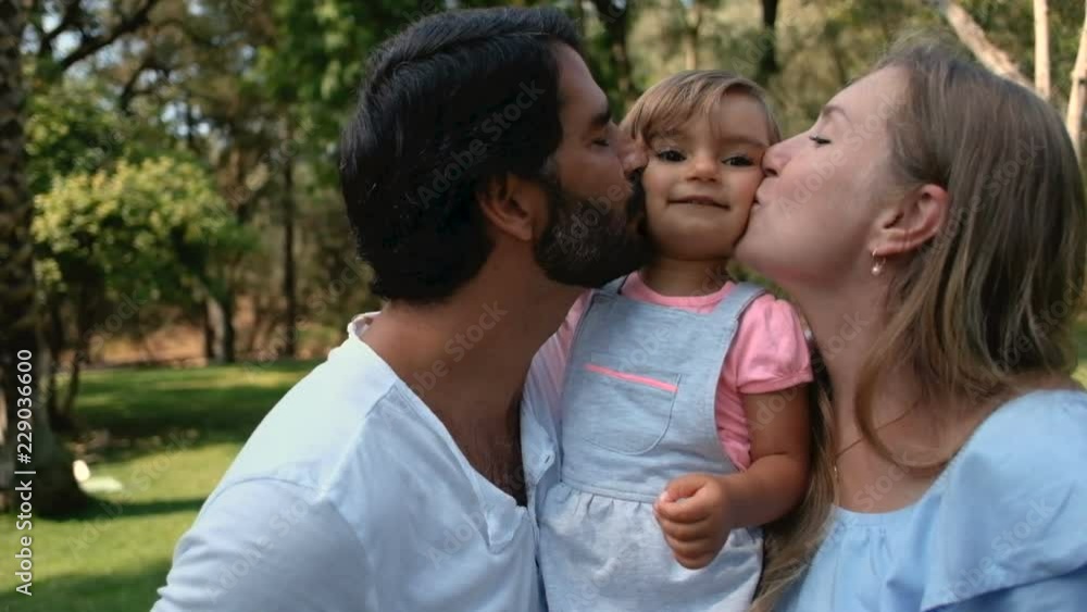Happy mother and father hugging and kissing little daughter in sunny park, front view. Cheerful parents kissing girl in slow motion. Family concept Happy mother and father hugging and kissing little daughter in sunny park, front view. Cheerful parents kissing girl in slow motion. Family concept