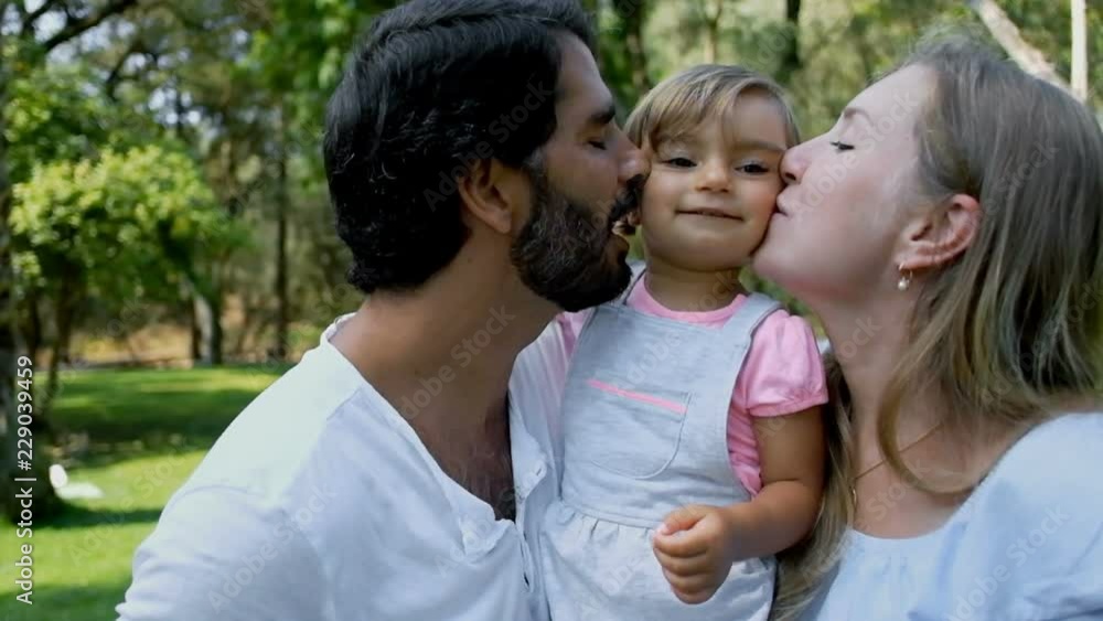 Smiling parents holding and kissing daughter in sunny park in slow motion. Cheerful mom and dad hugging little adorable girl, front view. Togetherness concept Smiling parents holding and kissing daughter in sunny park in slow motion. Cheerful mom and dad hugging little adorable girl, front view. Togetherness concept