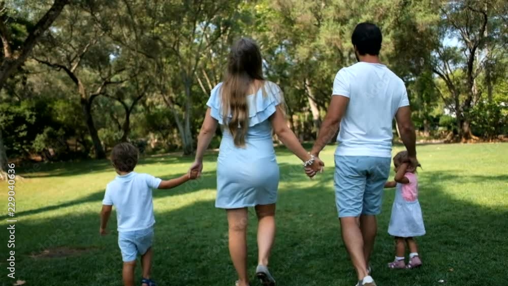 Young family holding hands with kids and walking in park together, back view. Happy father, mother and siblings walking on green lawn. Family walk concept Young family holding hands with kids and walking in park together, back view. Happy father, mother and siblings walking on green lawn. Family walk concept