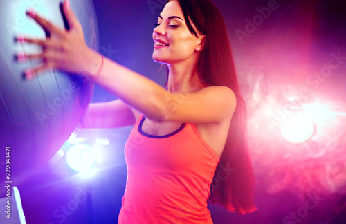Fitness girl yoga exercising in gym with ball. Happy woman with fitball works in multi-colored rays of sport room with lens flare. Neon flash light background.