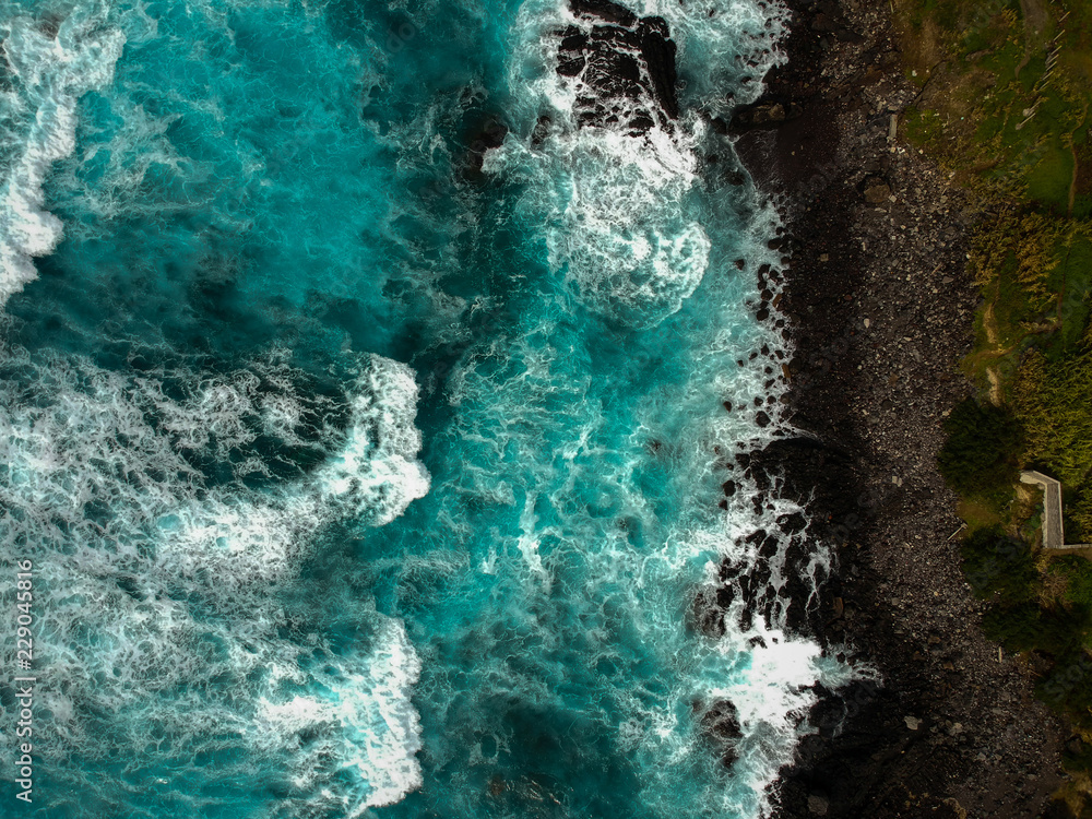 Aerial top view of sea waves hitting the coastline with black volcanic rocks with turquoise sea water. Seascape in the Portuguese coastline. Azores islands. Drone shot.
