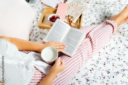 Young woman drinking milk at home in bed and reading book, top view