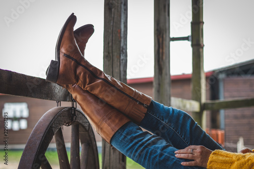 Woman is relaxing on the ranch with her legs on a wooden railing. Leather boot and jeans