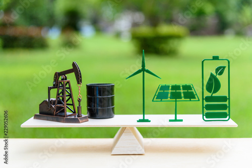 Fossil fuel vs renewable / future clean alternative energy concept : Petroleum pumpjack, crude oil drum barrel, solar panel, green leaf battery, wind turbine on a wood balance scale in equal position.