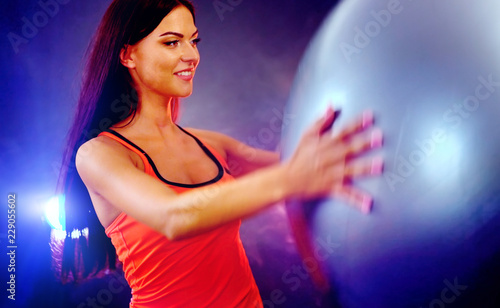 Fitness girl yoga exercising in gym with ball. Woman with fitball works in multi-colored rays of sport room. Portrait of full-length in profile with lens flare. Flash light background.