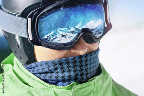 Close up of the ski goggles of a man with the reflection of snowed mountains. A mountain range reflected in the ski mask. Man on the background blue sky. Wearing ski glasses. Winter Sports. 