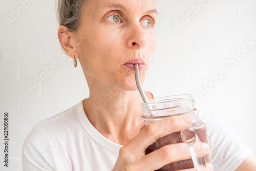 Closeup of middle aged woman drinking berry smoothie from glass mug with metal straw against white background (selective focus) photo