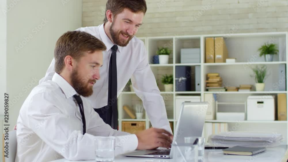 Medium shot of young twin brothers working in office together, one of them using laptop computer and one standing near him and discussing project Medium shot of young twin brothers working in office together, one of them using laptop computer and one standing near him and discussing project