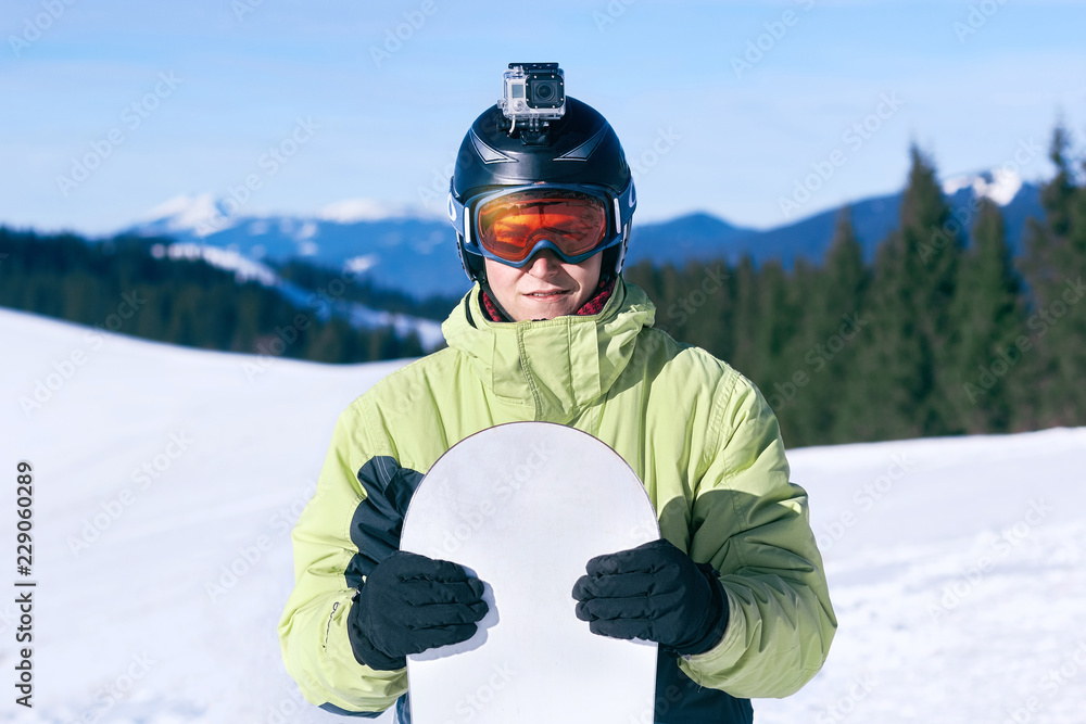 .Snowboarder with action camera on a helmet. Ski goggles  with the reflection of snowed mountains. Portrait of man at the ski resort on the background of mountains and blue sky,  hold snowboard. 