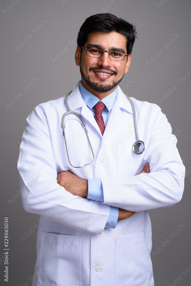 Young handsome bearded Persian man doctor against gray backgroun Young handsome bearded Persian man doctor against gray backgroun