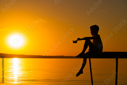 a little boy sits on the masonry with a paper boat in his hand at sunset near the river. Silhouette of a boy on the background of the evening landscape