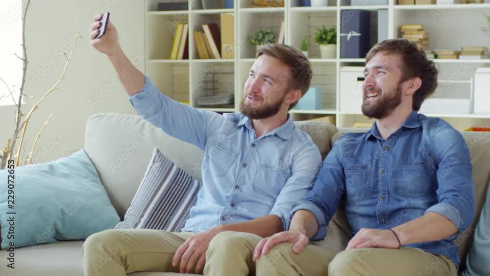 Handheld shot of two cheerful bearded twin brothers sitting on sofa together and laughing while taking selfie on smartphone Handheld shot of two cheerful bearded twin brothers sitting on sofa together and laughing while taking selfie on smartphone