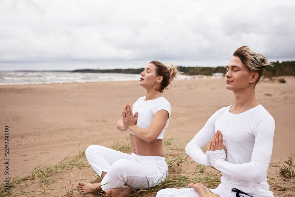 Two atheltic European people man and woman in white sportswear closing eyes  and holding hands in namste gesture, sitting in padmasana while meditating  outdoors during yoga retreat by the ocean Stock Photo |