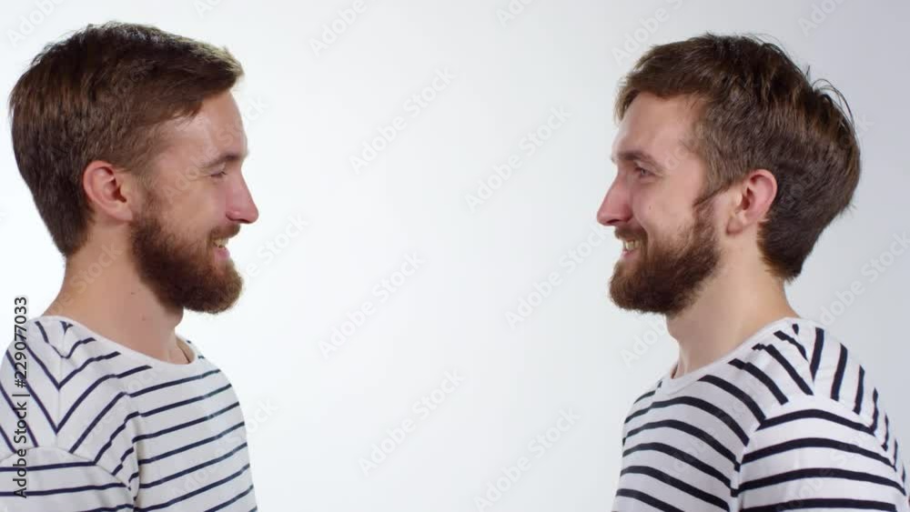 Medium shot of two young bearded twin brothers standing opposite each other on white background, looking each other in the eye and laughing Medium shot of two young bearded twin brothers standing opposite each other on white background, looking each other in the eye and laughing